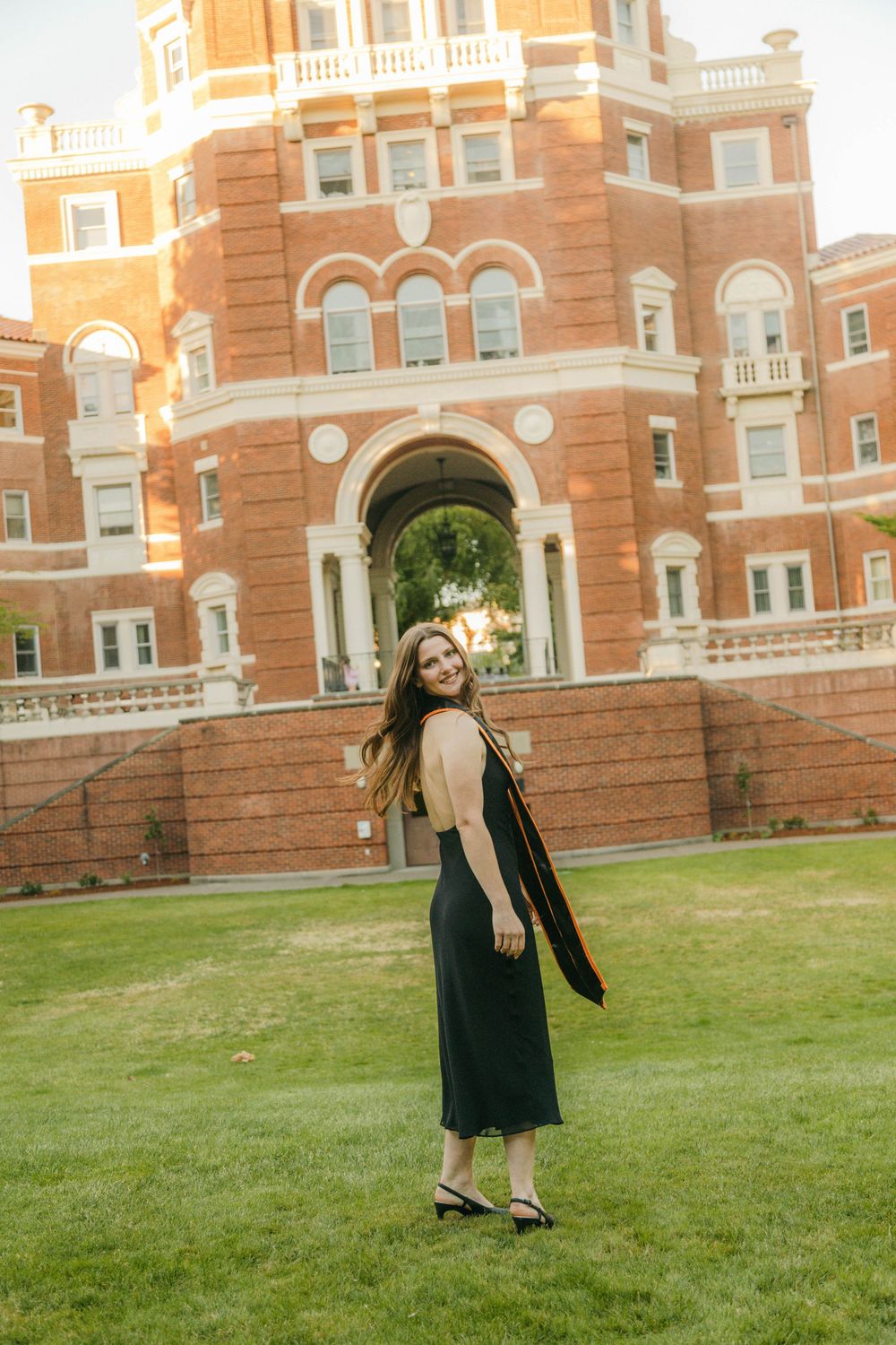 A smiling graduate in a black dress holds her cap outside a red brick university building on a sunny day.