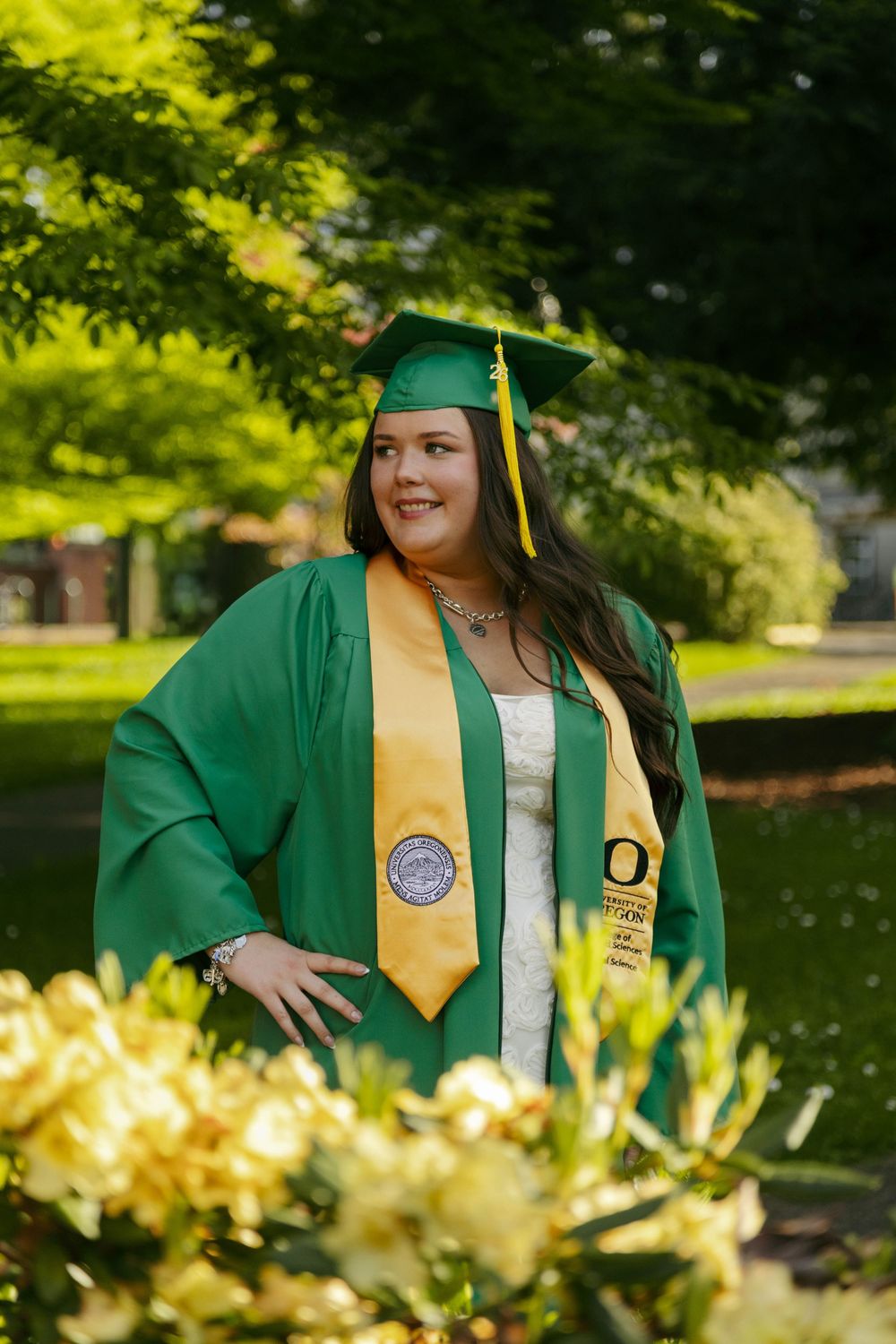 A smiling graduate poses outdoors in a green cap and gown with a gold stole, surrounded by lush greenery and yellow flowers.