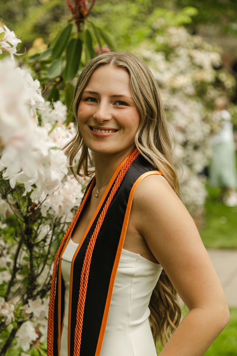 Smiling female graduate wearing black and orange stole poses beside white blooming flowers in lush green garden.