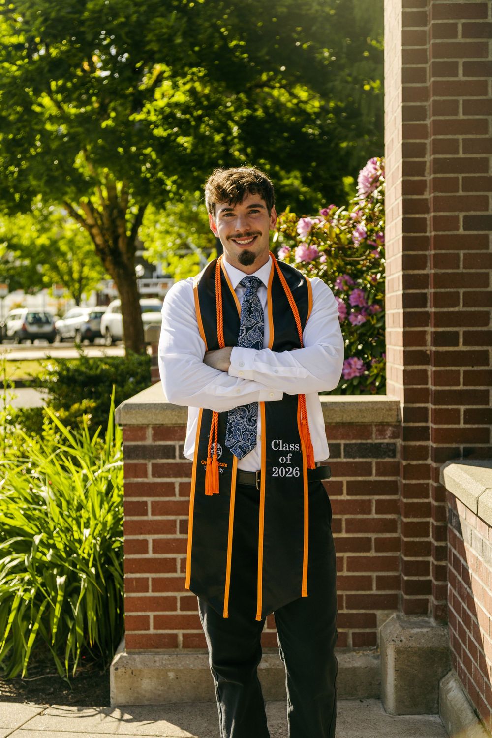 Young man in graduation stole reading Class of 2026 poses arms crossed outdoors near brick building and blooming flowers.