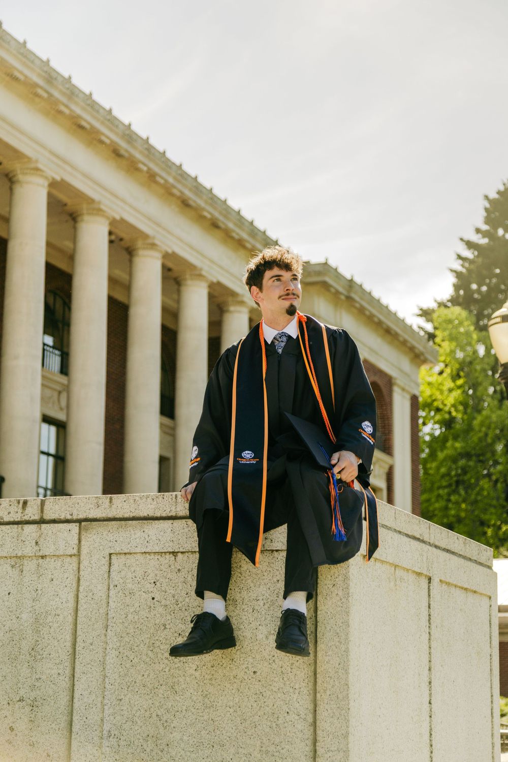 A young man in black graduation gown with orange sash sits on stone steps before a columned university building.