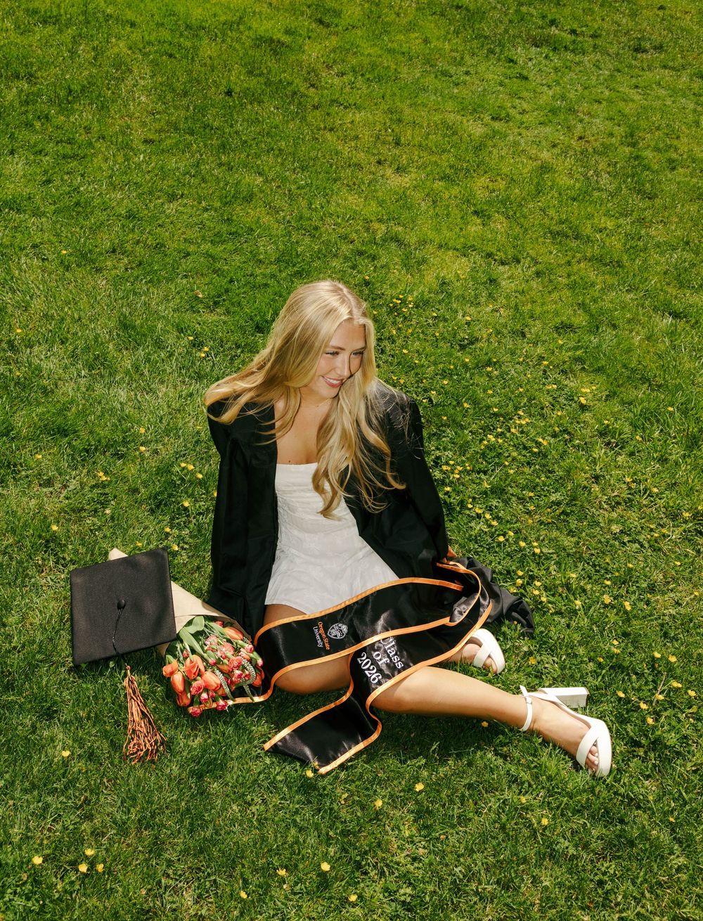 Blonde woman in black blazer sitting on grass with graduation cap, sash, and flower bouquet for senior photos.
