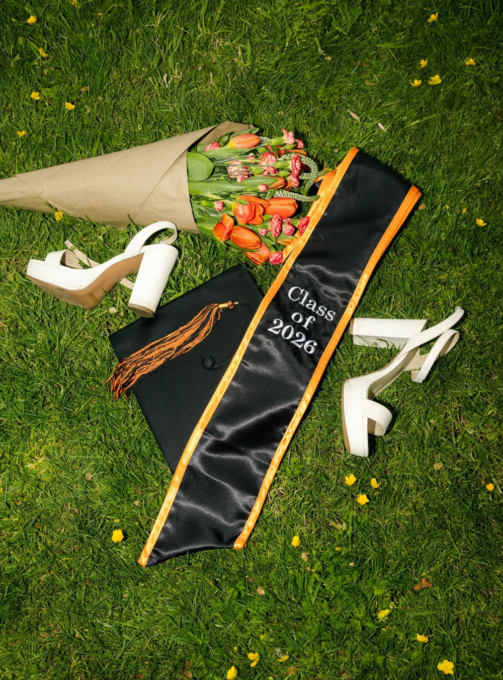 Class of 2026 black and orange graduation sash with cap, white heels, and tulip bouquet on green grass.