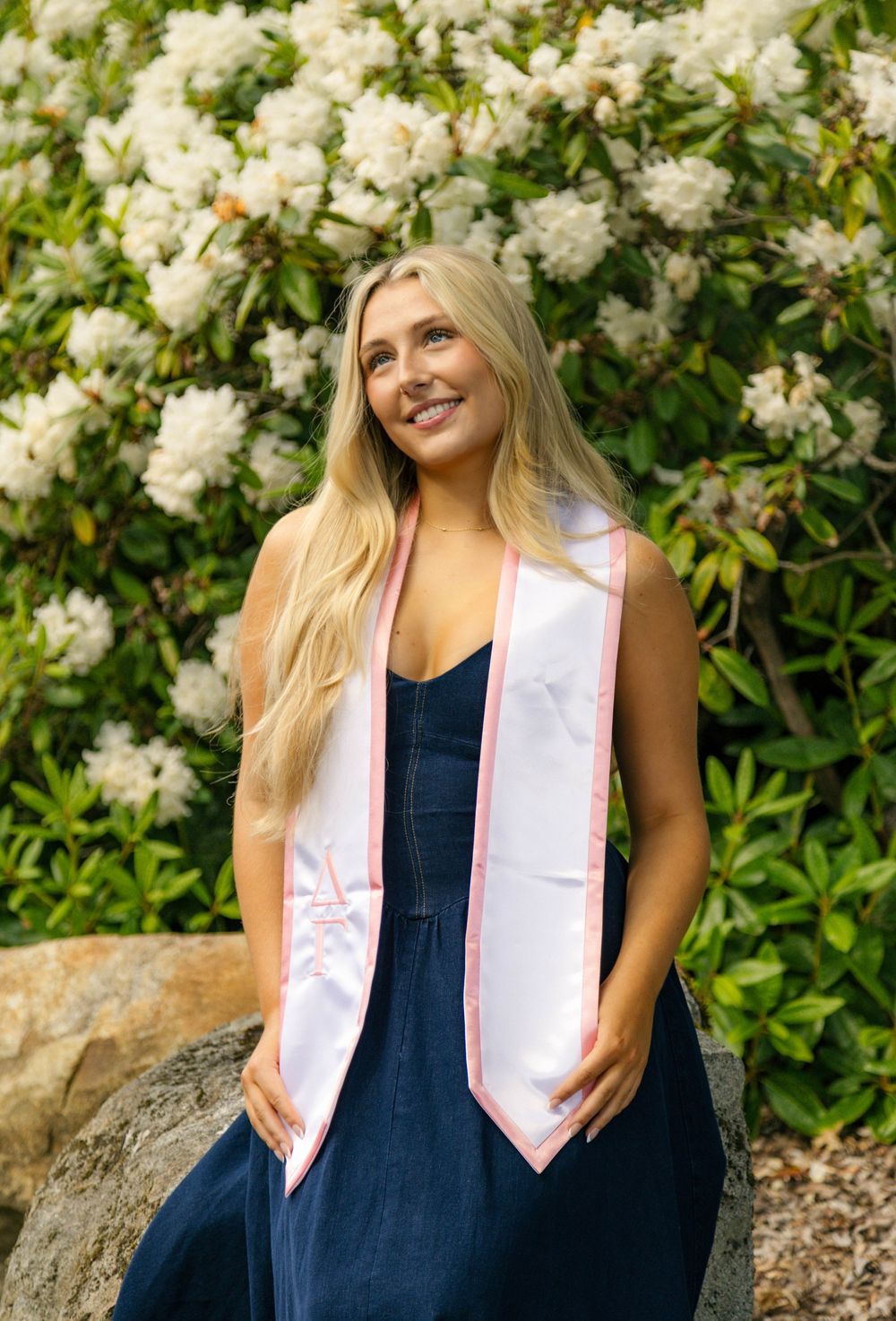 Smiling blonde woman wearing a white and pink graduation stole seated in front of blooming white flowers.