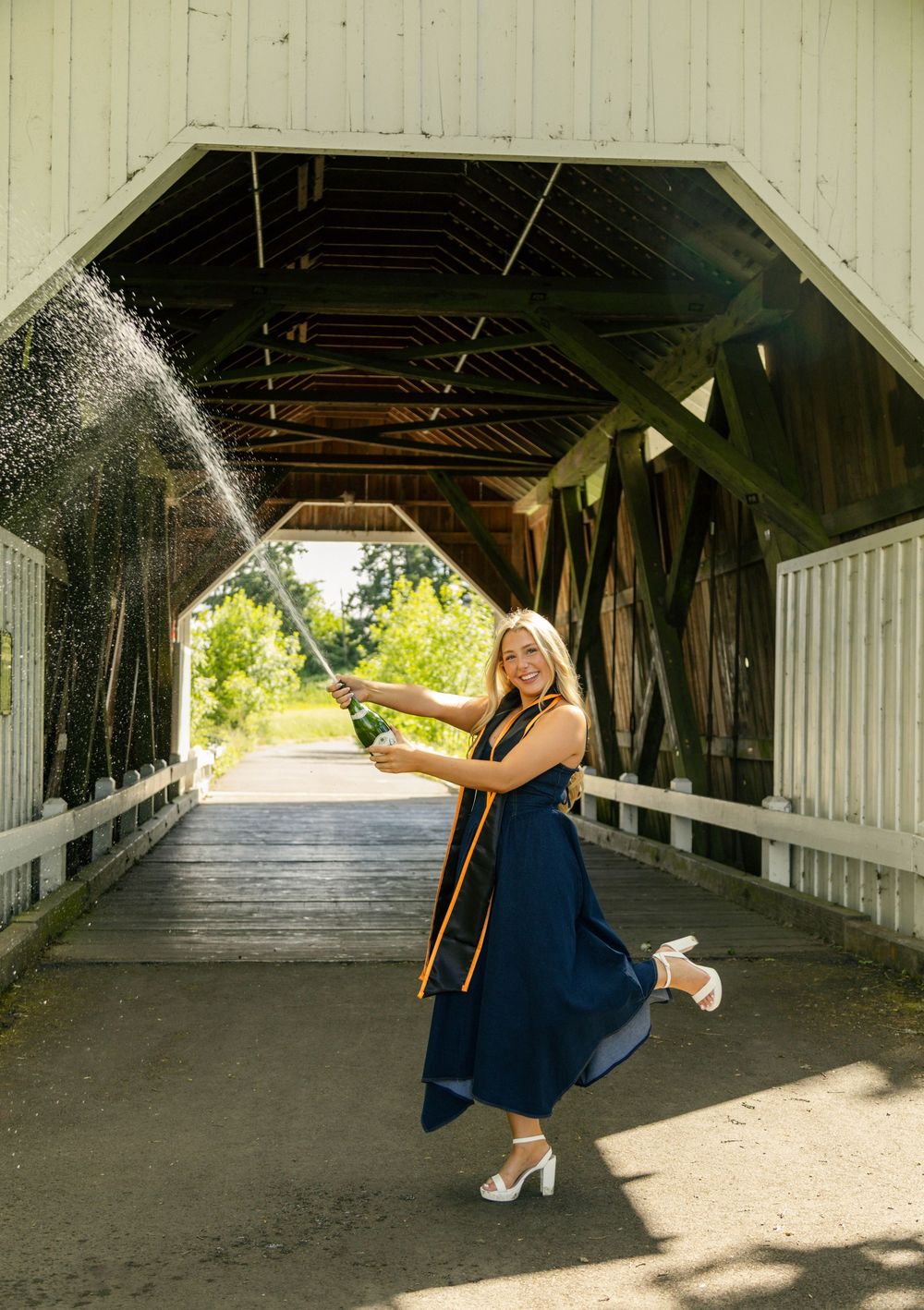 Smiling graduate in navy dress sprays champagne under a covered wooden bridge during an outdoor celebration.