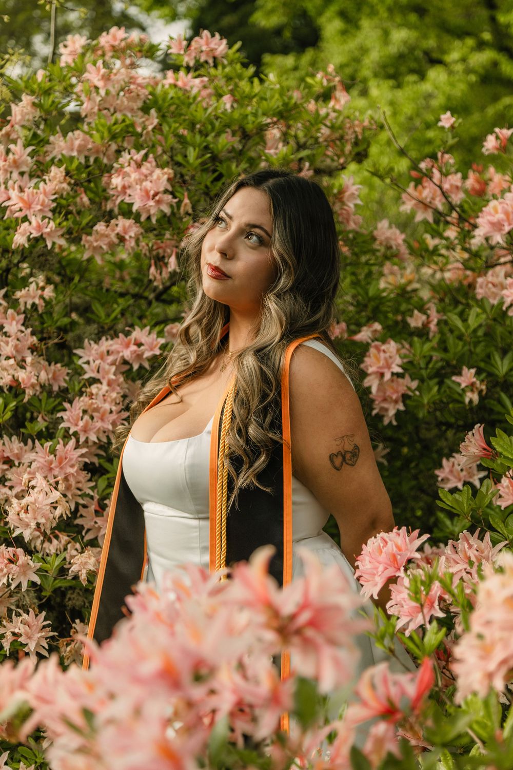 A young woman in a white dress and orange graduation sash poses among blooming pink azalea bushes.