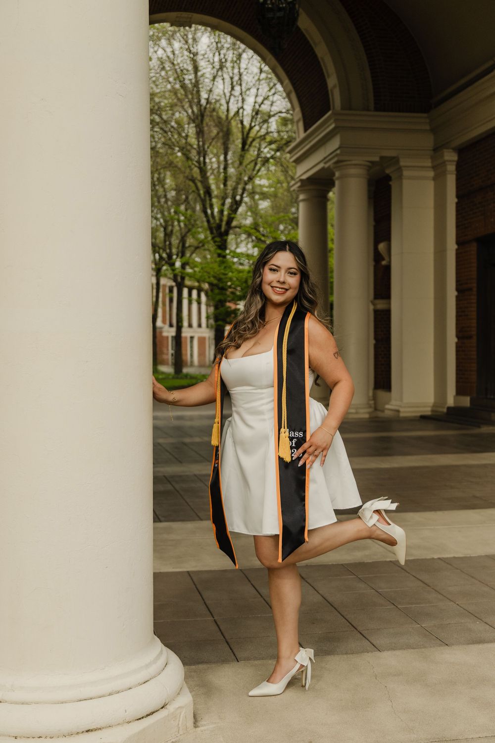 Smiling graduate in white dress and stole poses by column outdoors.