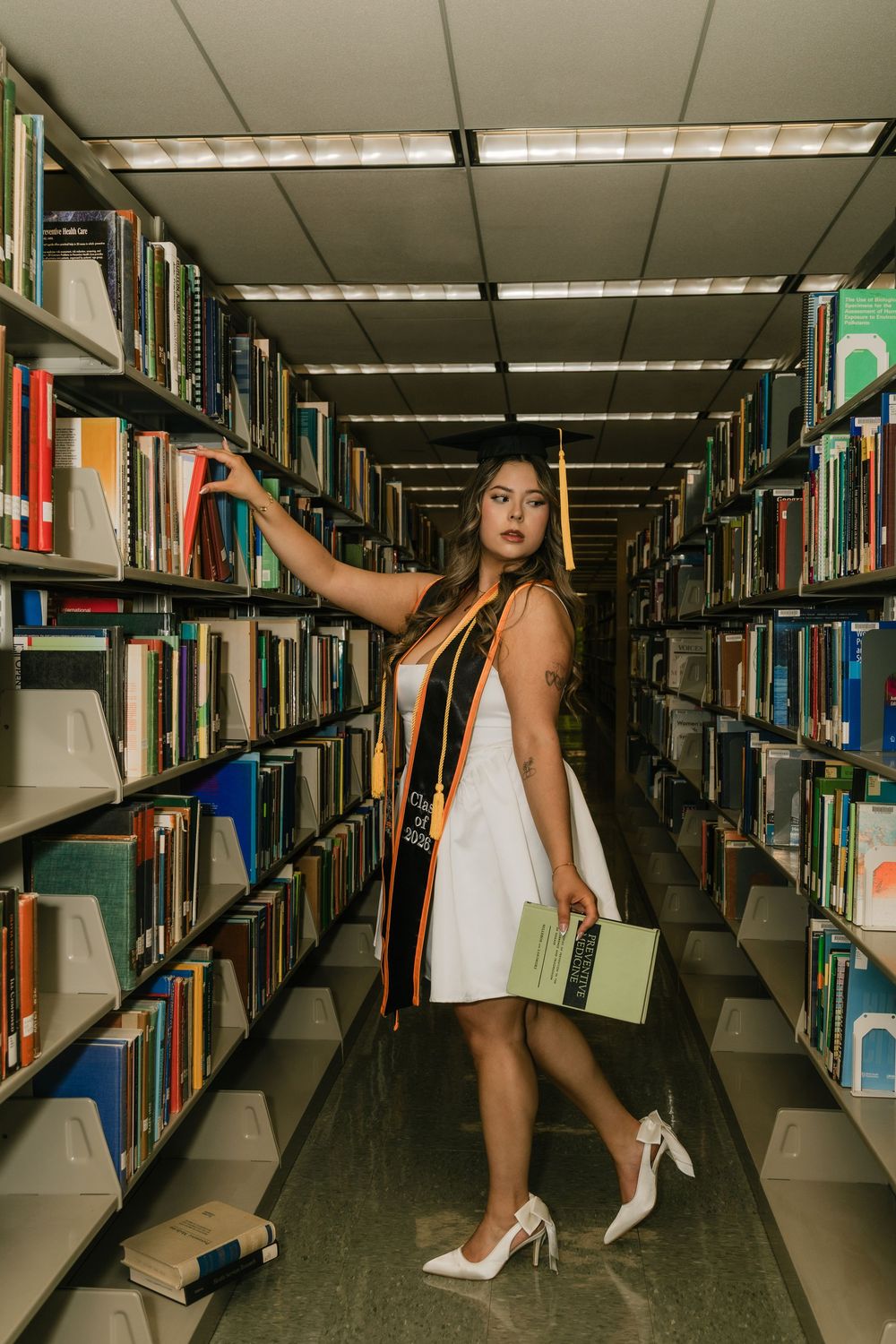Graduate in white dress and stole holds book between library shelves.