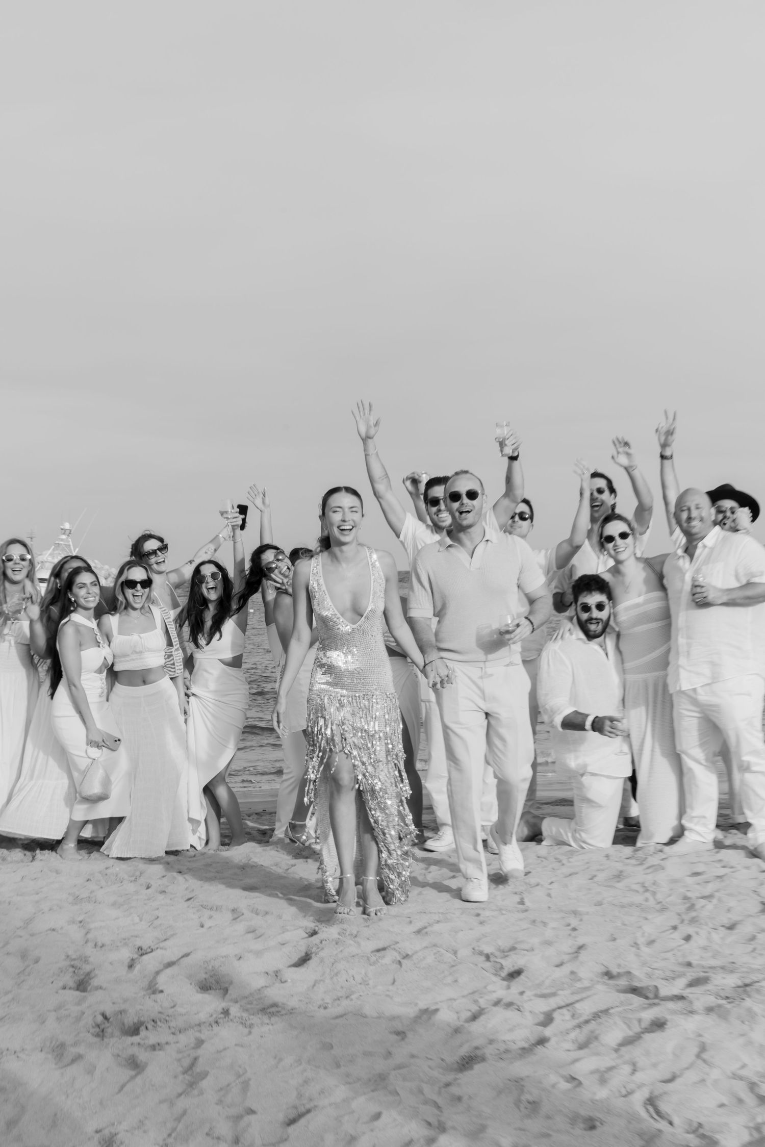 A celebratory group in white attire jumps joyfully on a sandy beach in black and white.