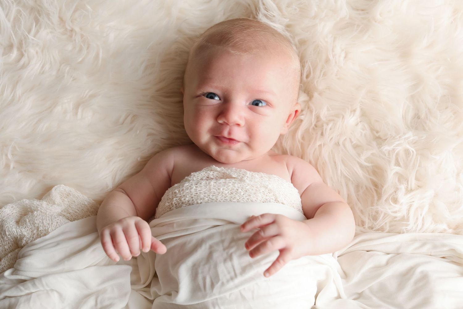Baby wrapped in cream fabric lying on soft white textured background.
