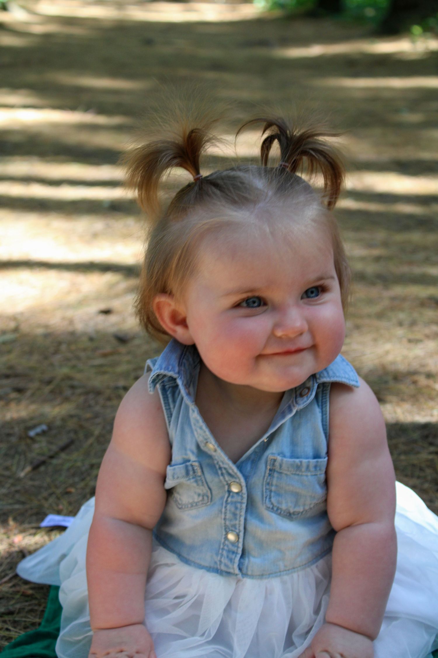 Baby wearing denim and white dress sitting outdoors in dappled sunlight.