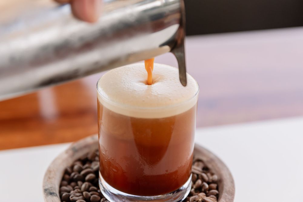 Layered espresso drink being poured into a glass with coffee beans on the base.