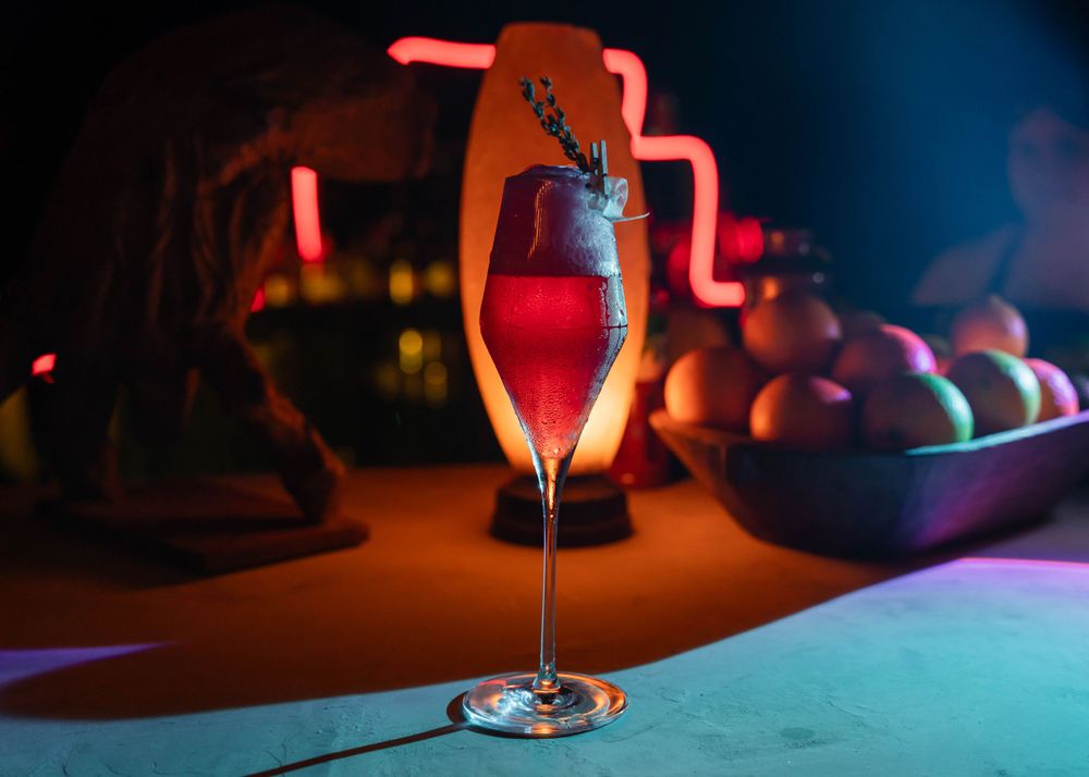 Red cocktail in champagne flute with herb garnish against neon-lit background.