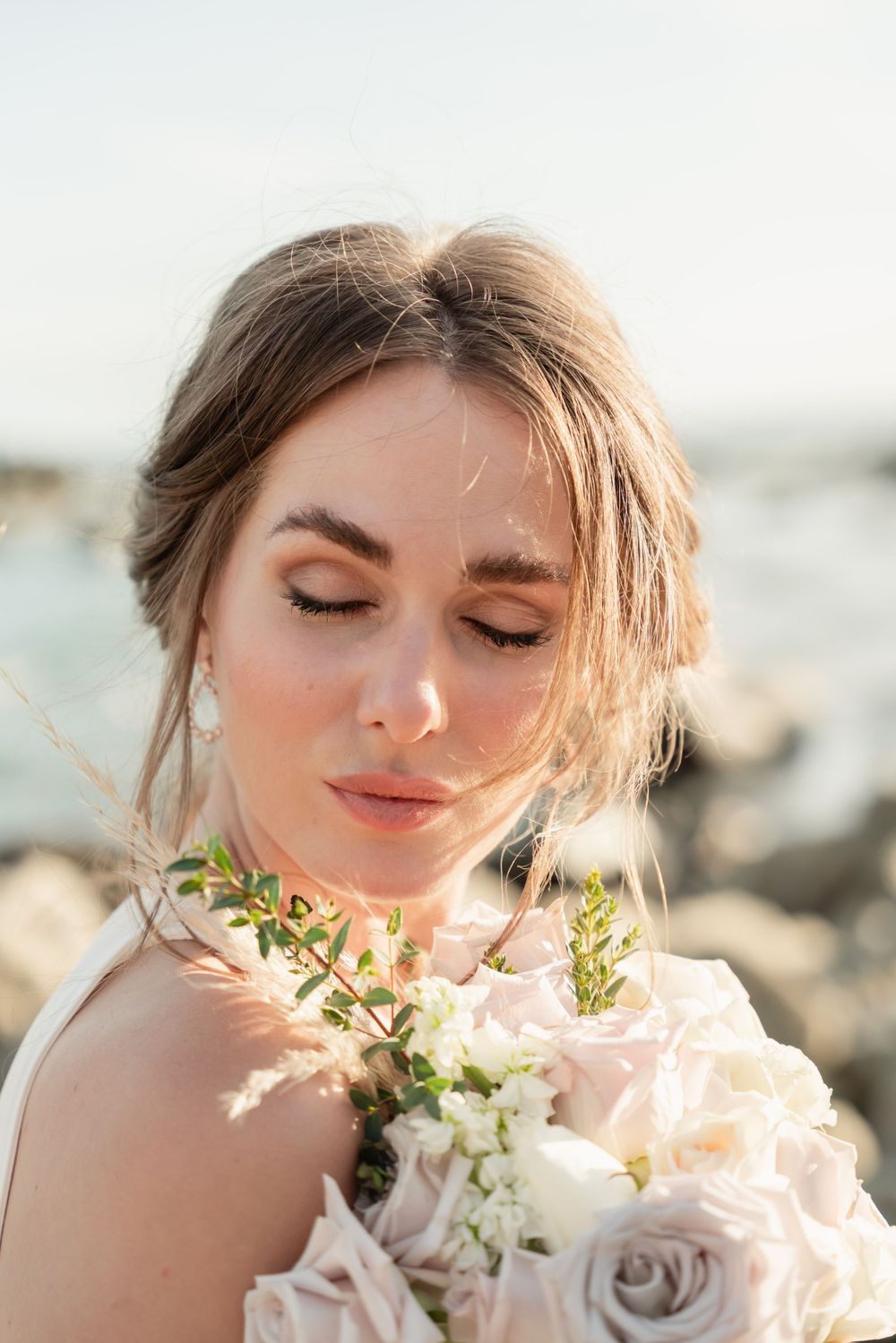 Ethereal bridal portrait with delicate pink and white roses against a coastal backdrop at sunset.