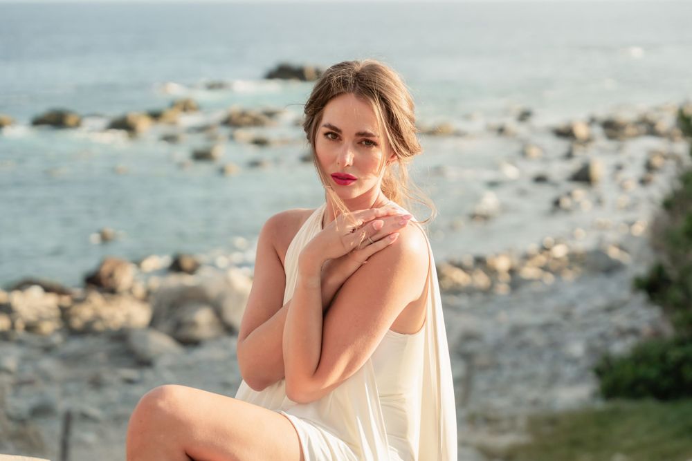 Elegant seaside portrait in a flowing white dress against rocky coastal backdrop.