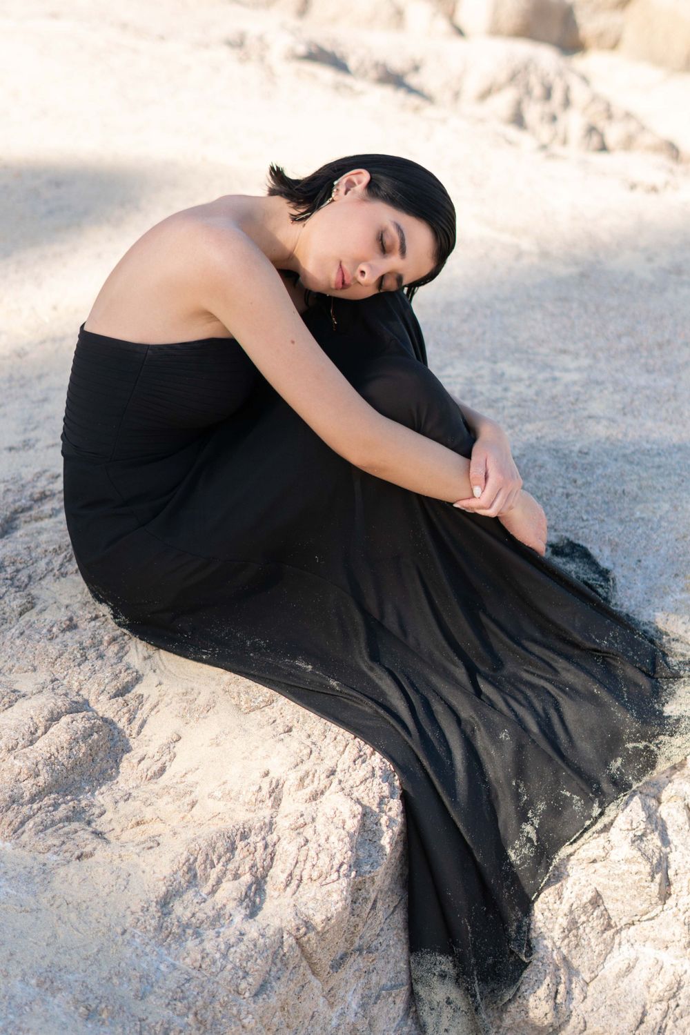 A figure in a flowing black dress poses gracefully on coastal rocks at sunset.