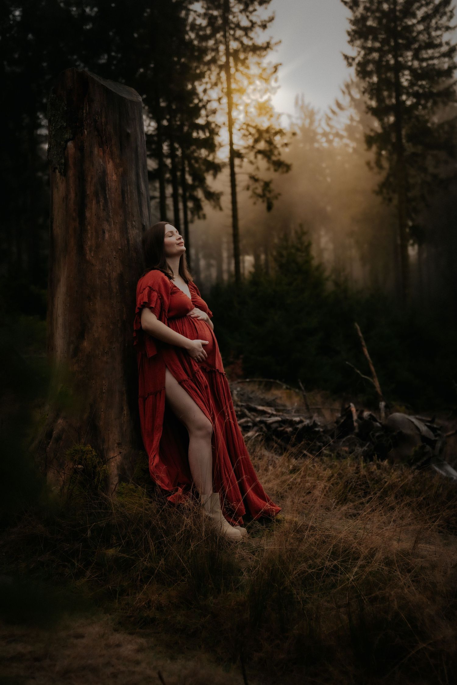 A flowing red dress creates a dramatic silhouette against a misty forest backdrop at sunset with tall pine trees.