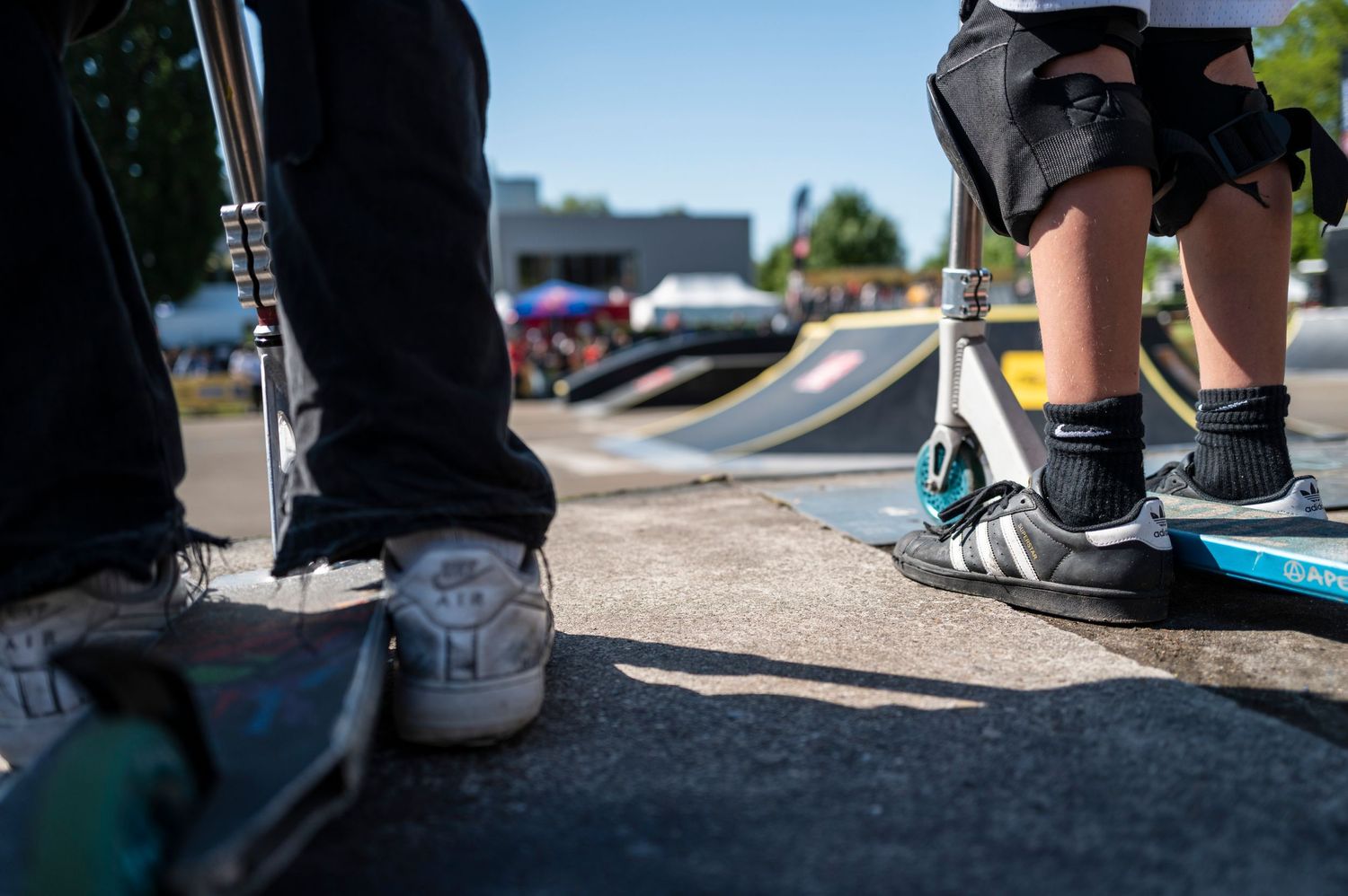 Shoes and feet of skateboarders standing on concrete at a skate park.