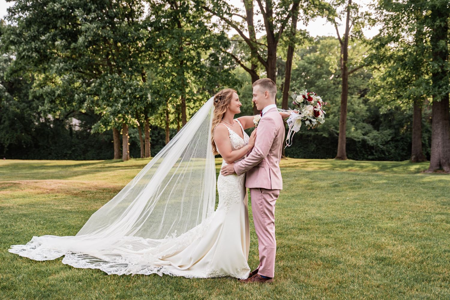 Bride & groom sharing a joyful moment outdoors, with the bride in a white gown & groom in a blue tuxedo, smiling at each other.