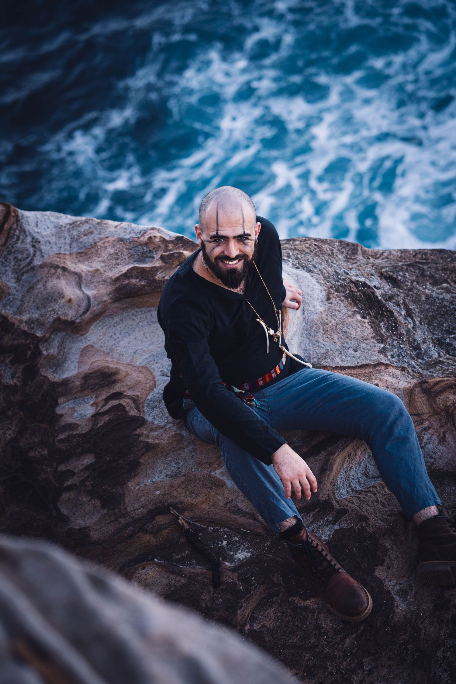 A viking themed male portrait on coastal rocks shows someone in black sweater and jeans above crashing waves.