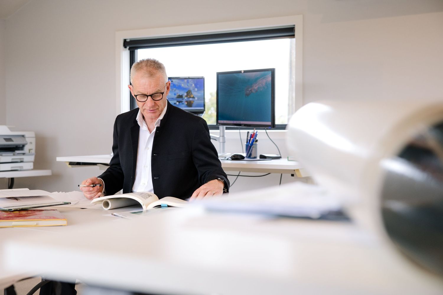 Professional reviews paperwork at desk with window and computer monitors in background.