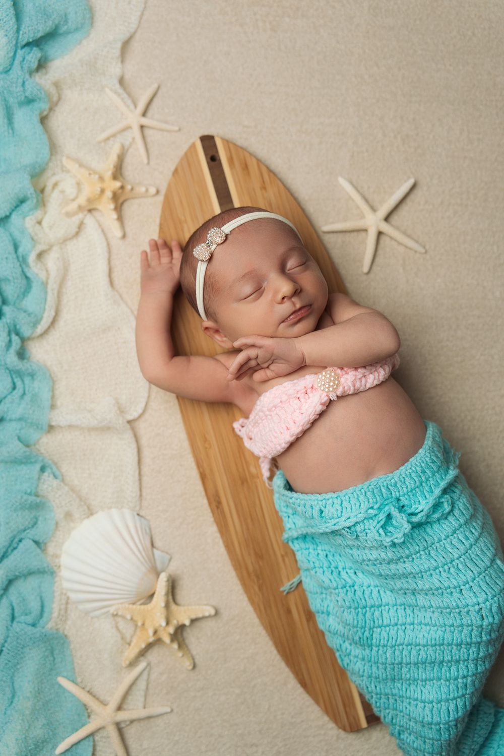 Newborn baby in turquoise knit wrap posed with seashells and starfish on a surf board in a beige backdrop in beach-themed photo series.
