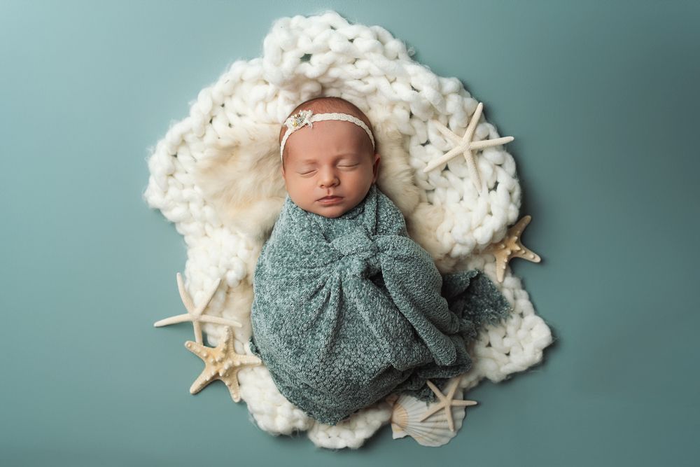 A sleeping newborn baby wrapped in a teal swaddle blanket lies on white textured fabric with soft teal backdrop.
