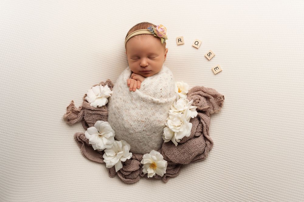 A newborn baby wearing white sleeps peacefully on a brown blanket adorned with delicate white flowers.