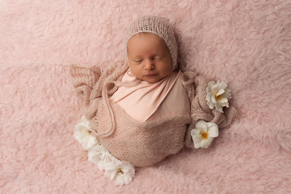 Sleeping newborn wrapped in pink fabric on textured rose background with white flowers.