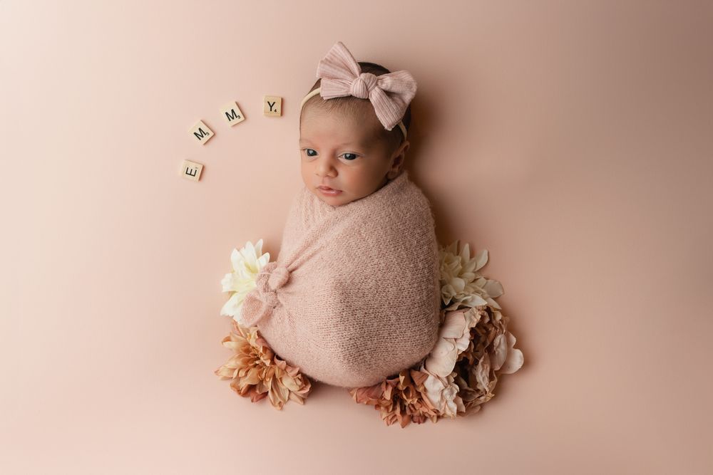 A newborn swaddled in soft pink fabric lies on a pink backdrop surrounded by delicate dried flowers and name letters.