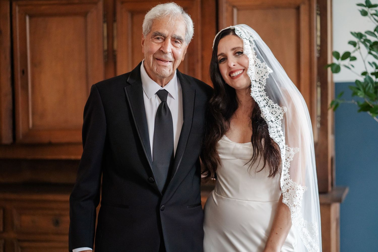 An elderly gentleman in a black suit stands with a bride wearing a white dress and lace veil.