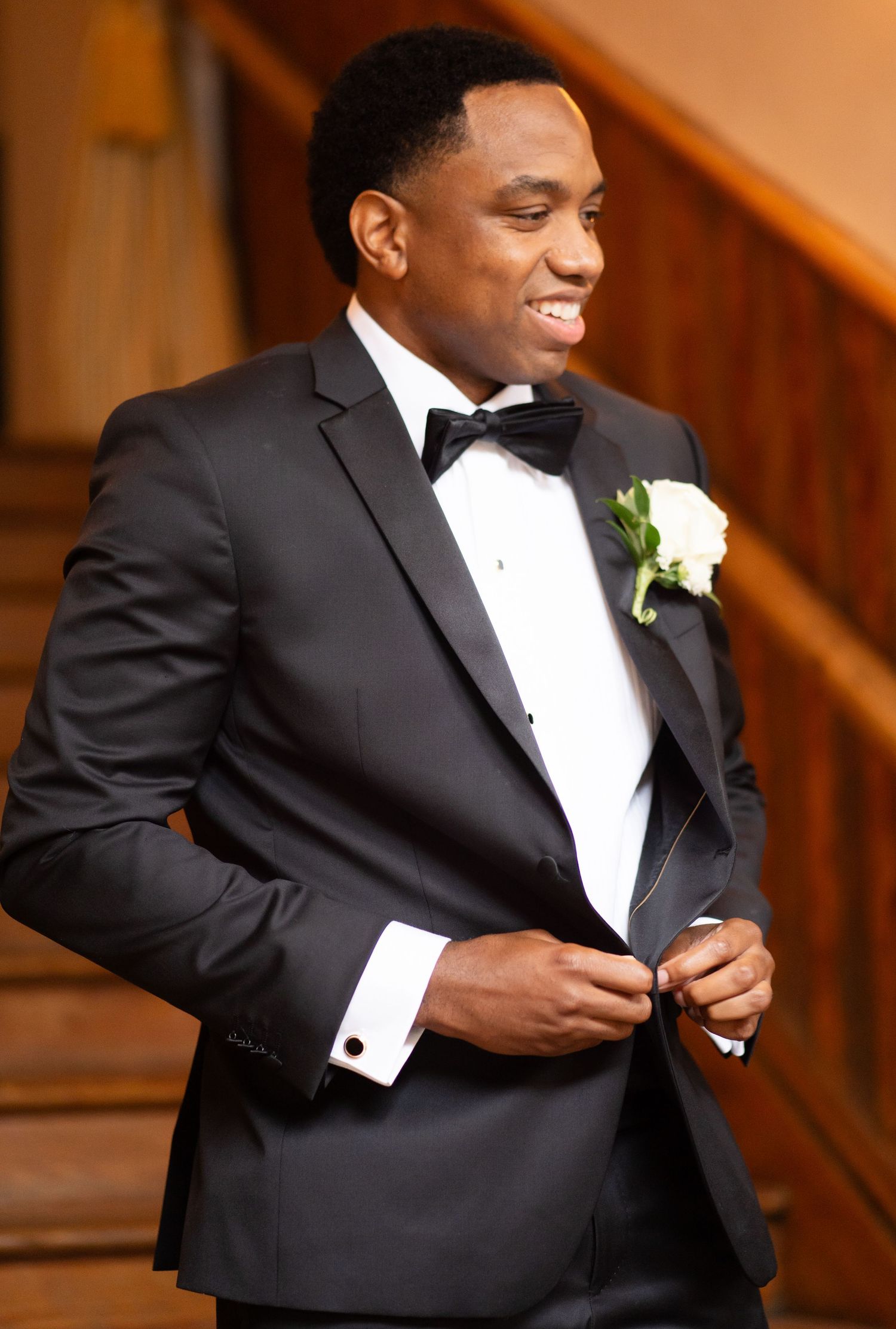 A well-dressed groom in a classic black tuxedo and bowtie smiles while adjusting his suit, with a white boutonniere.