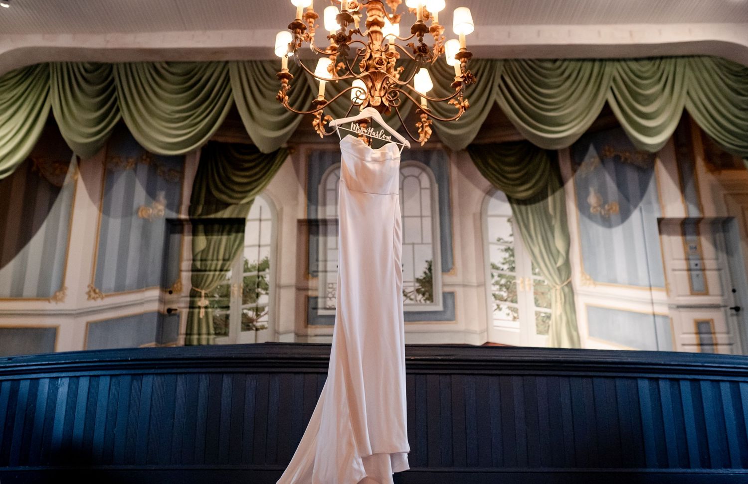 White wedding dress hangs elegantly beneath an ornate chandelier with green and blue draped curtains in background.
