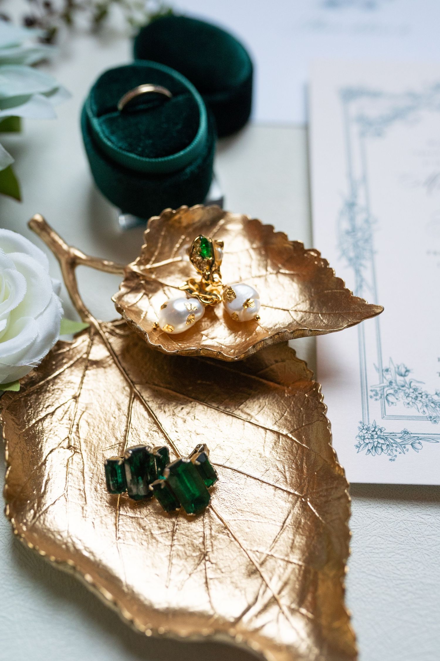 Wedding jewelry and earrings displayed on a decorative gold leaf dish with a dark green ring box against a white surface.