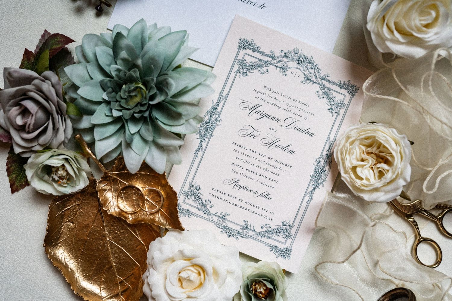 A wedding invitation surrounded by white roses, succulents, and gold accents on a light background.