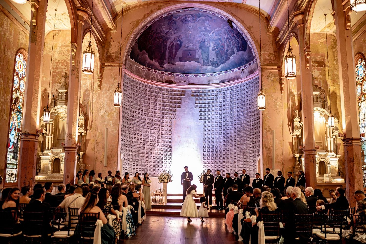 Wedding ceremony in an ornate church with domed ceiling, stained glass windows and dramatic purple lighting.