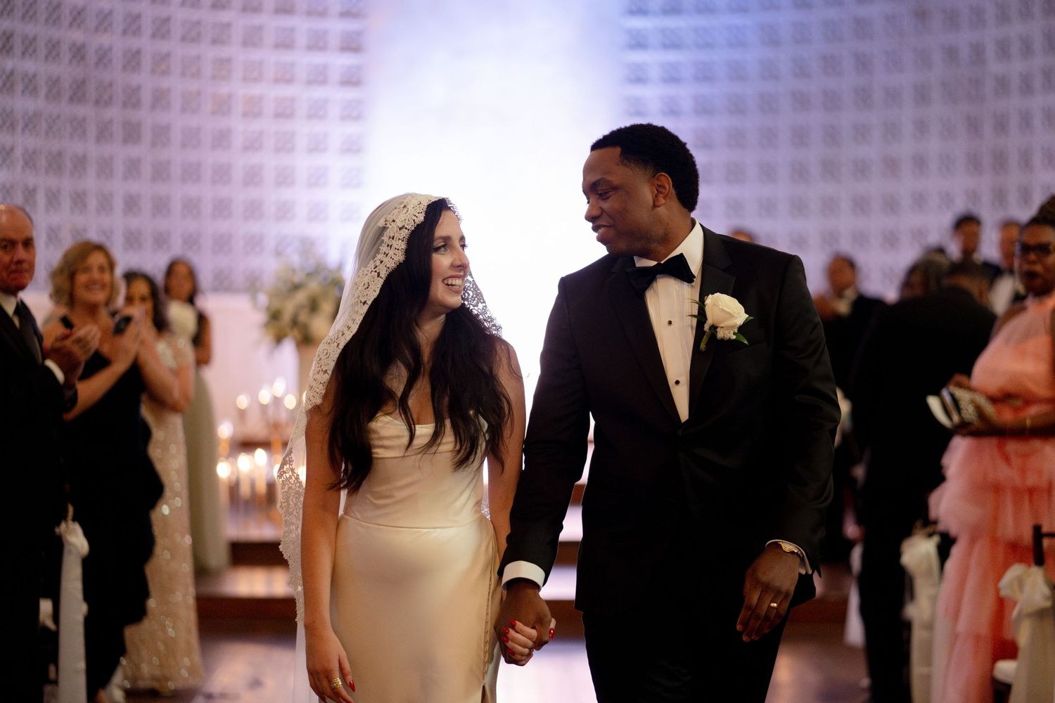 A bride and groom walk together holding hands during their white modern wedding reception.