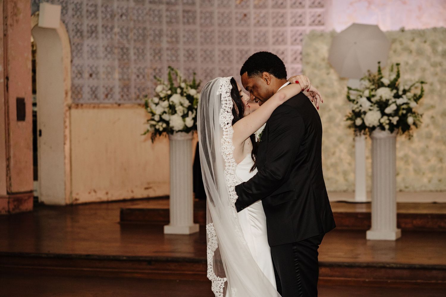 Two people share an intimate embrace at the altar of an elegant chapel decorated with white floral arrangements.