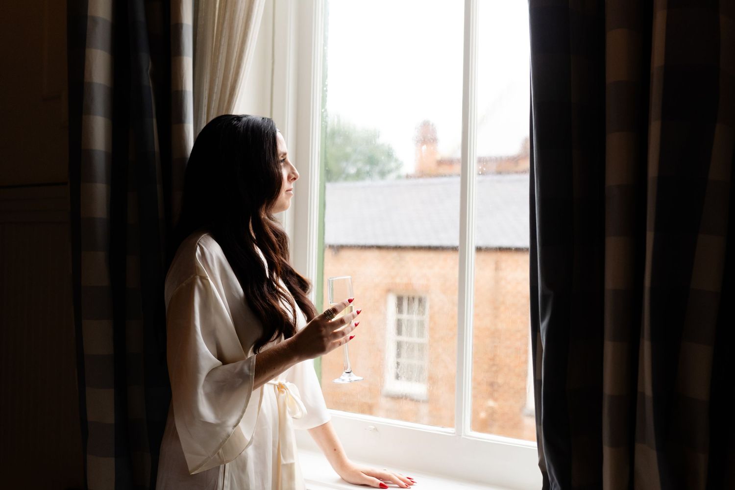 Person in white robe stands by window holding glass while looking out at urban view through dark curtains.