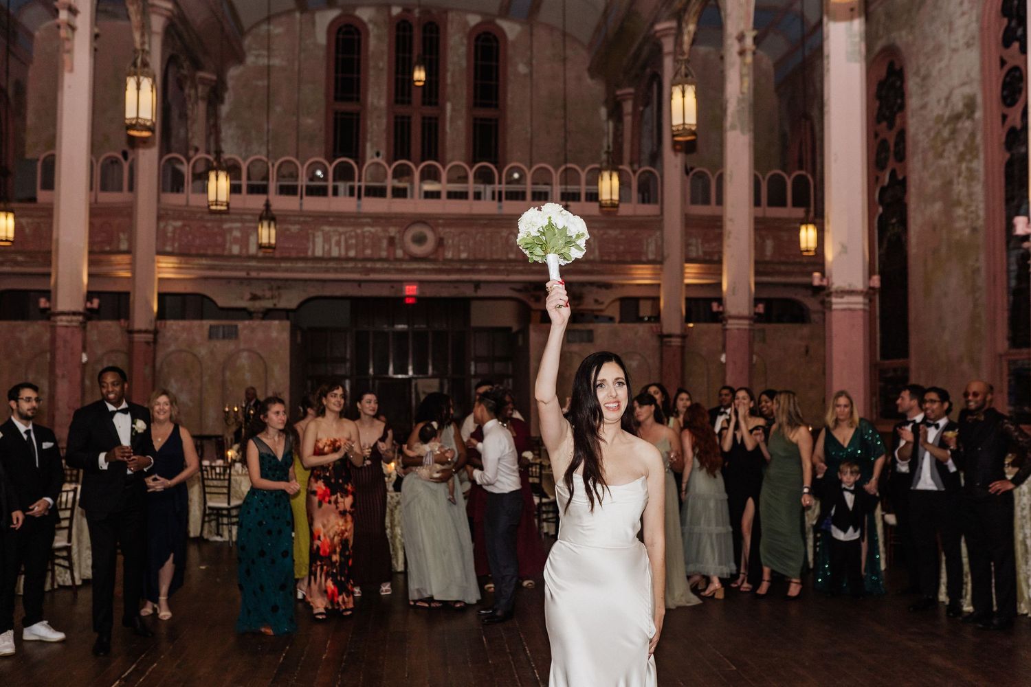 Wedding guest toss tradition in a grand historic ballroom with ornate columns and decorative lighting.