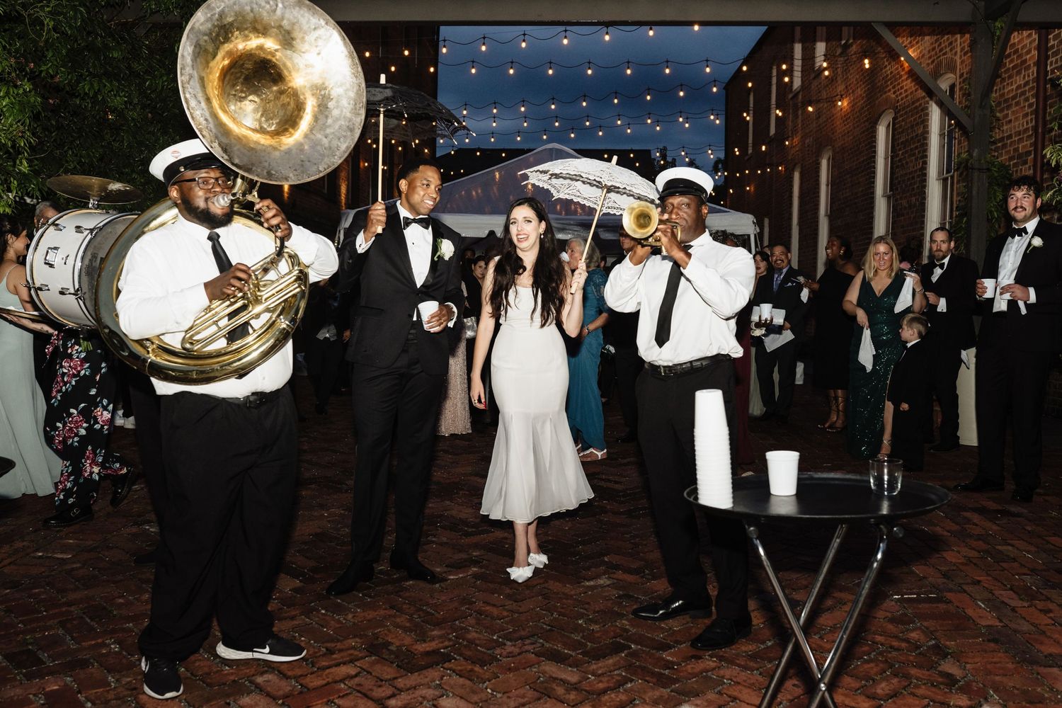 A lively wedding celebration with musicians playing brass instruments and guests dancing on a brick patio at night.