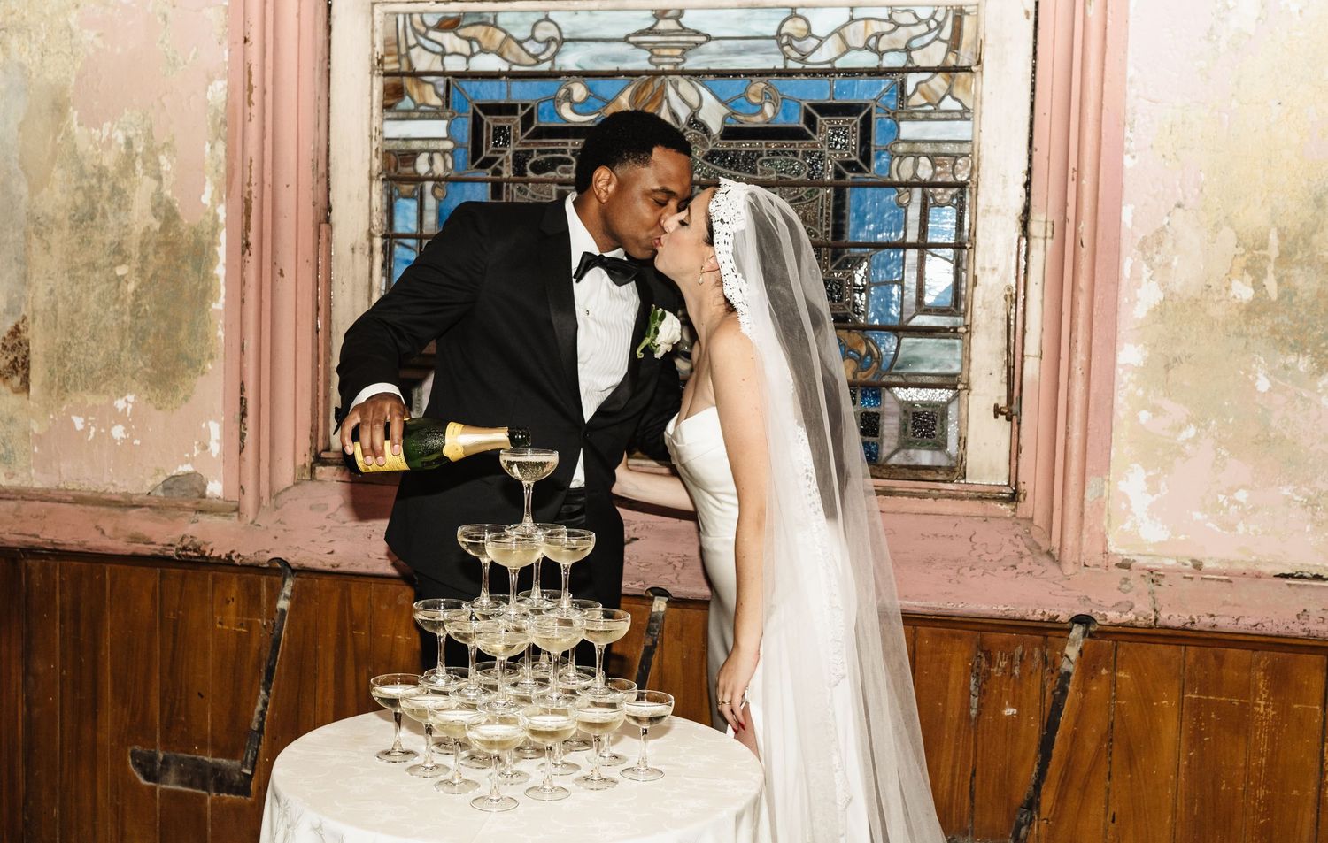 A couple in formal wedding attire pours champagne into a tower of champagne glasses at their reception.