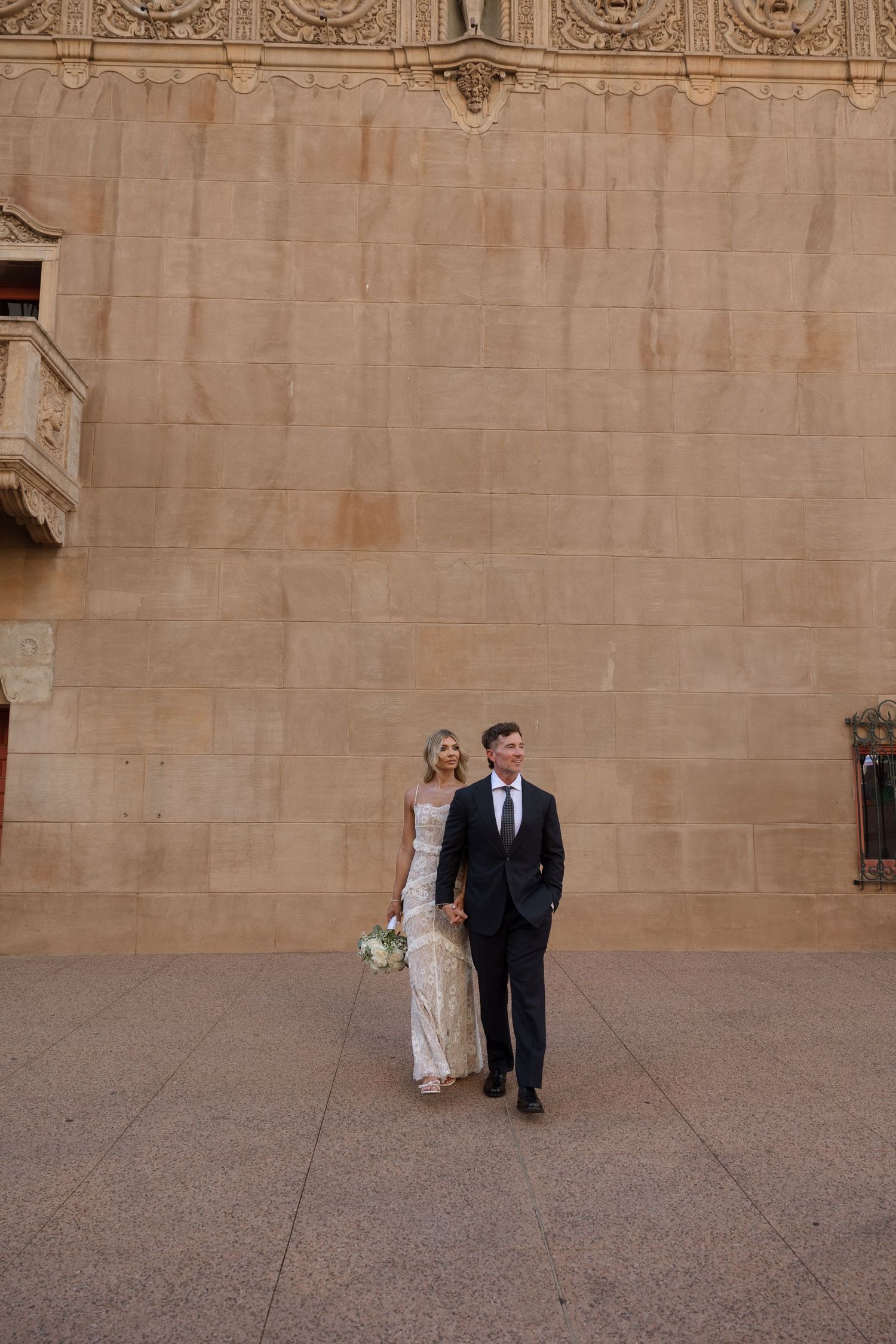 A couple in formal attire descend stone steps outside an elegant building with arched doorway and decorative lanterns.