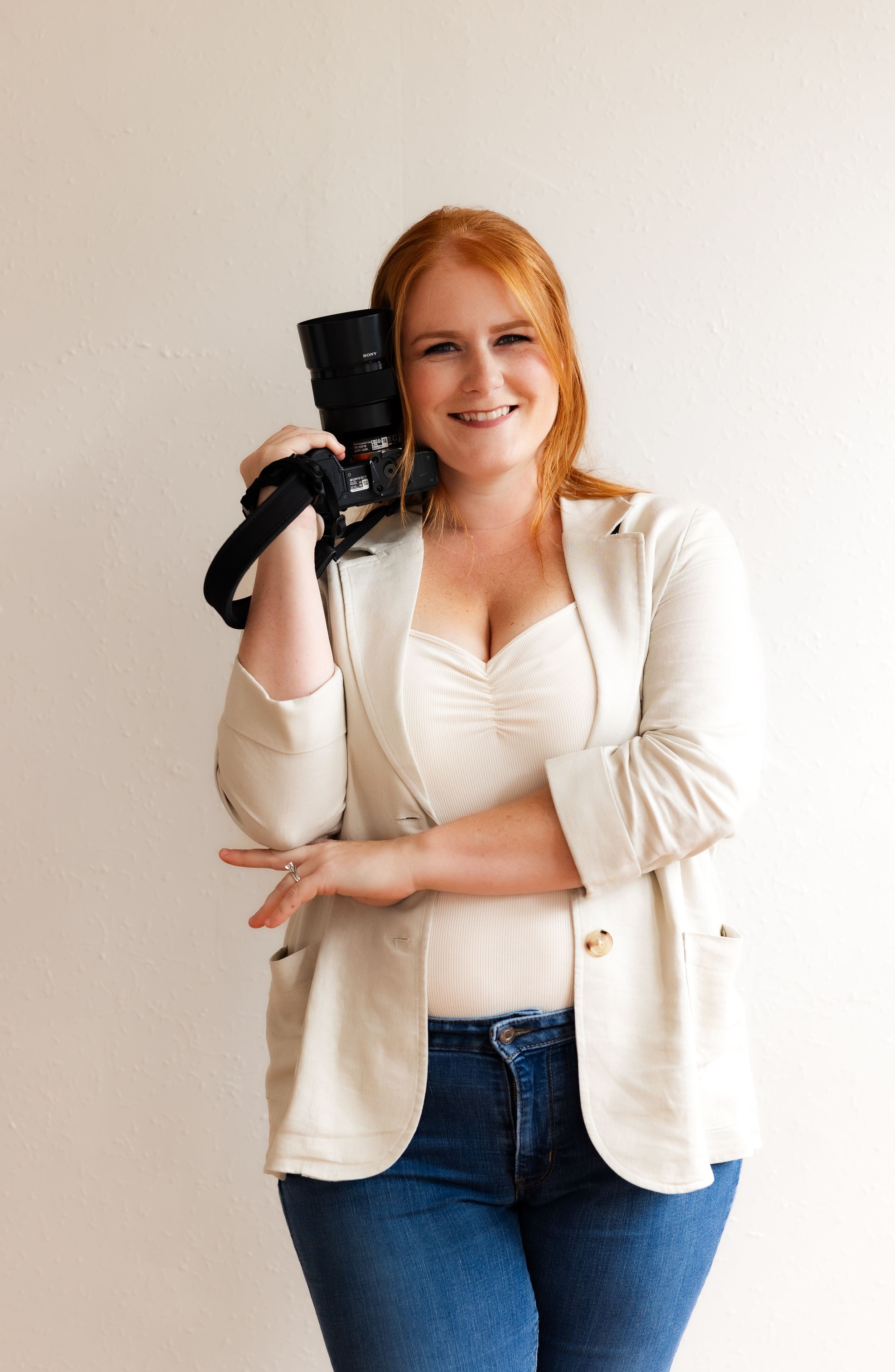 Professional photographer in white blazer and jeans holding a camera against white background.