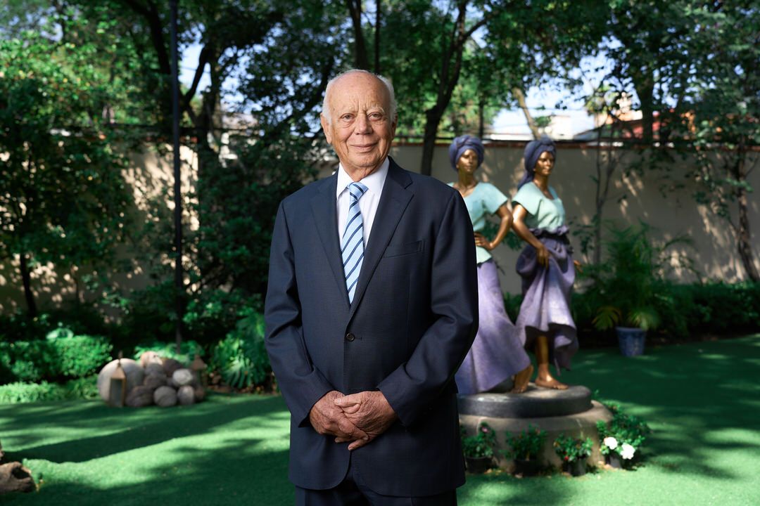 Retrato formal de socio senior o notario en jardín privado con esculturas y luz natural.