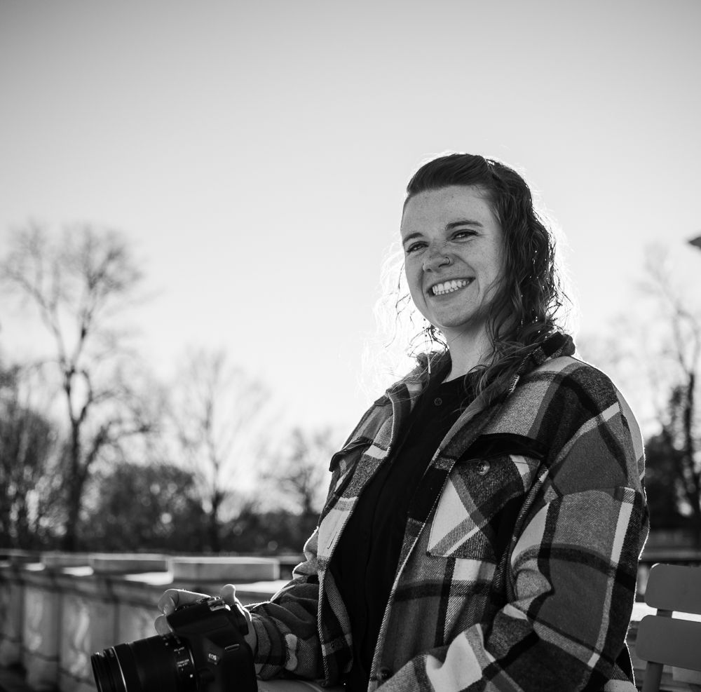 Smiling young woman holding a camera outdoors in black and white photo.