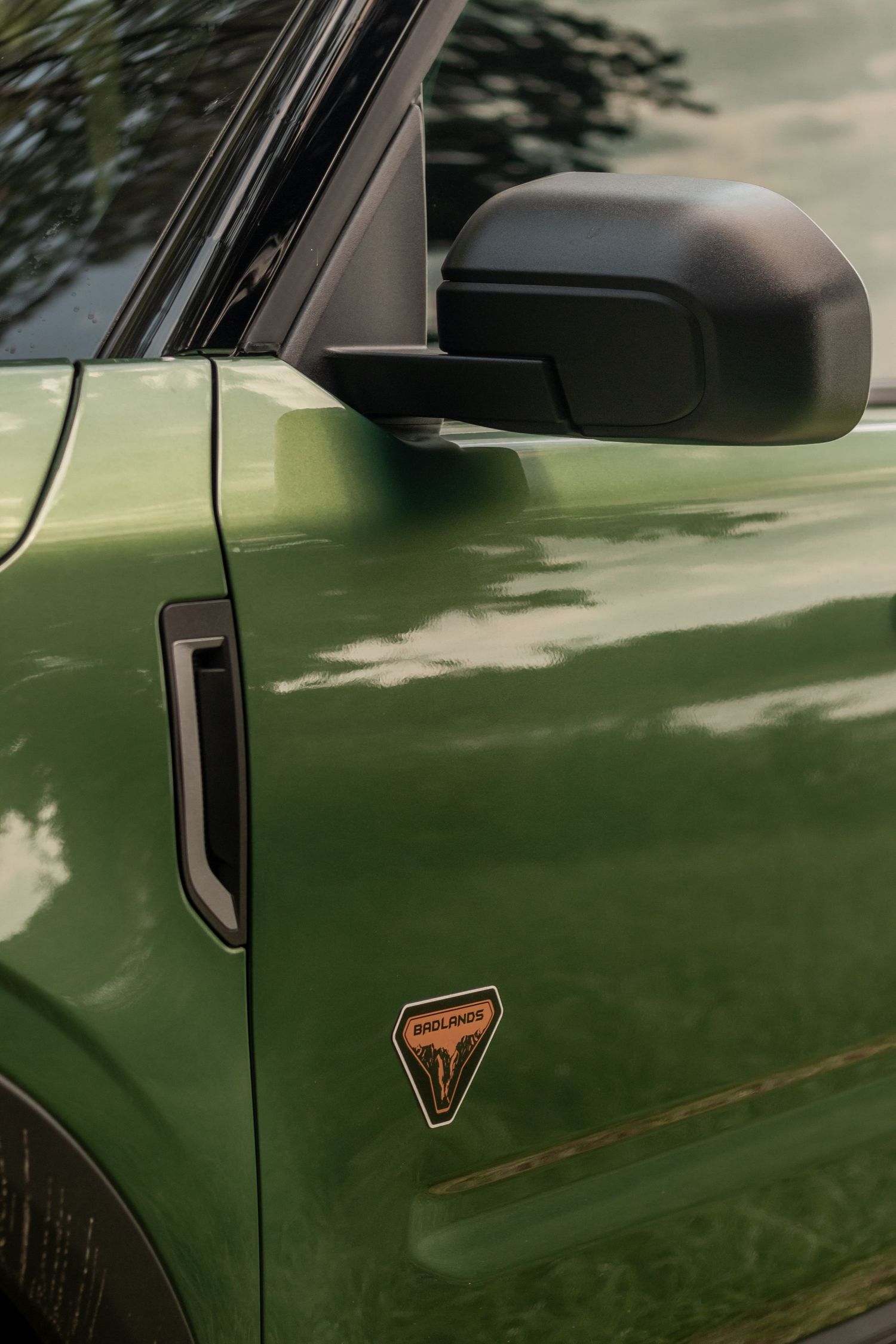 Close-up detail of green Ford Bronco Sport showing side mirror and badging against forest backdrop.