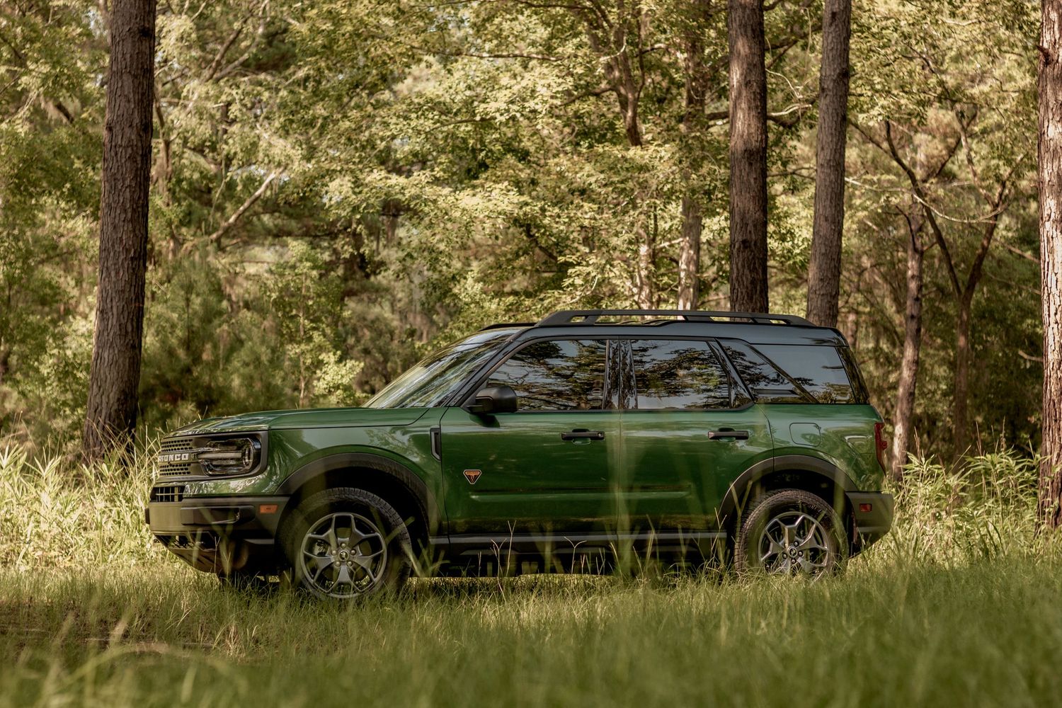 Green Ford Bronco Sport parked in a scenic wooded area with dappled sunlight filtering through the trees.