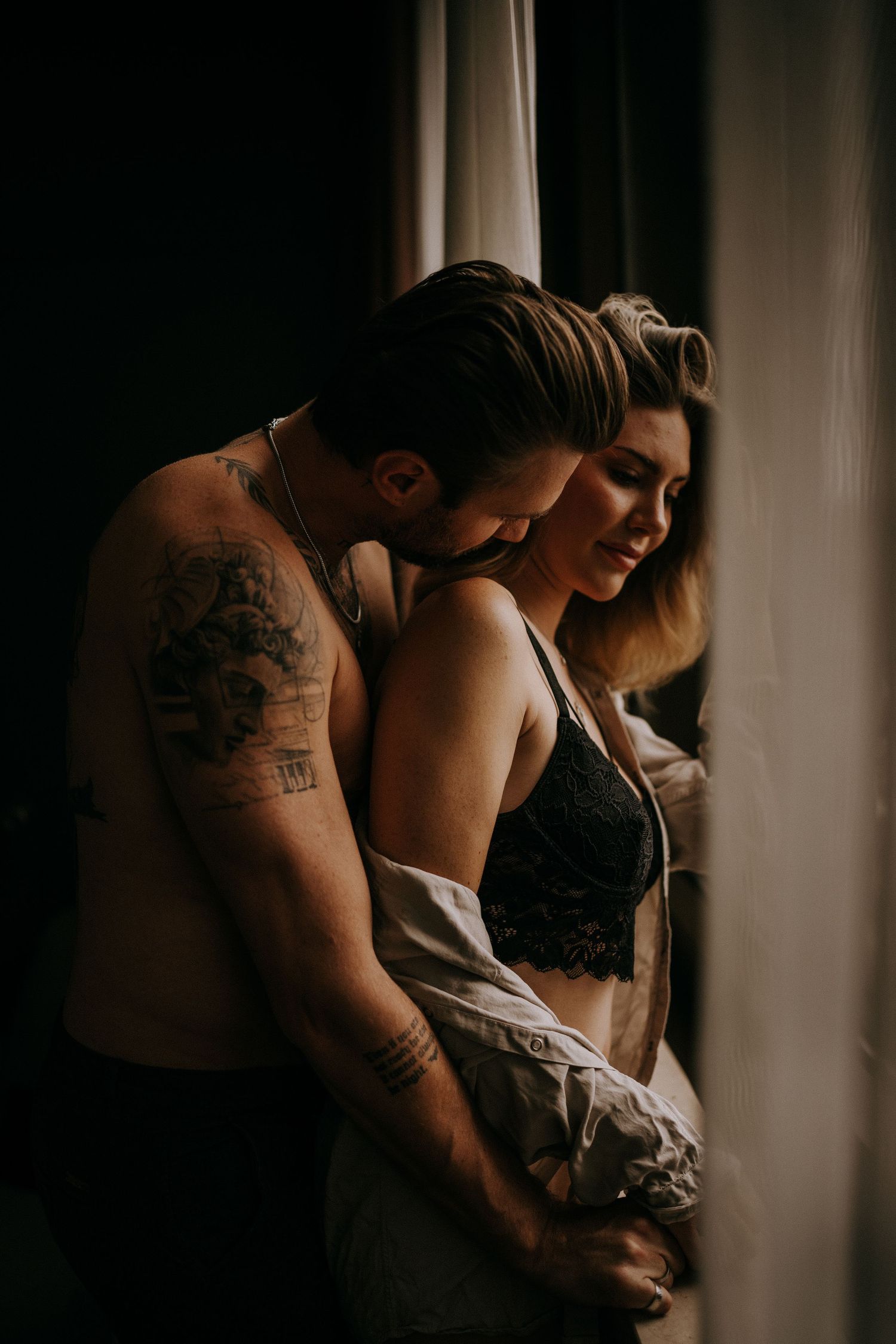 Intimate couple portrait with tattoos in moody window lighting.