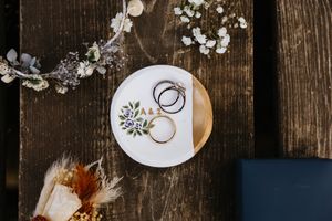 A flatlay of two wedding rings on an ornate plate surrounded by a flower arrangement on a rustic wood table taken at a Sacramento wedding.